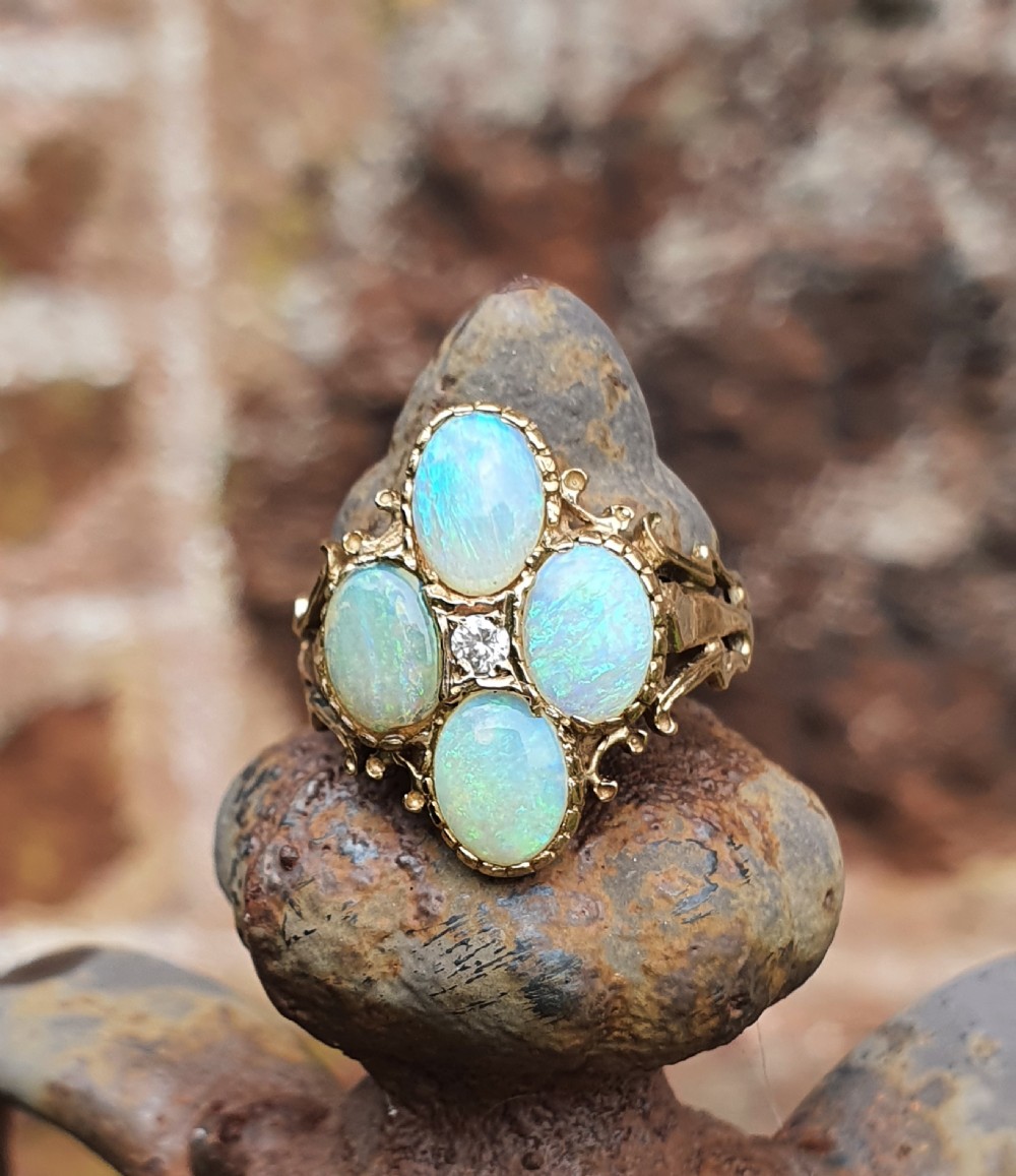 A 9ct yellow gold dress ring featuring four oval cabochon opals arranged in a cluster around a central brilliant-cut diamond, mounted on an ornate scrollwork band. View X