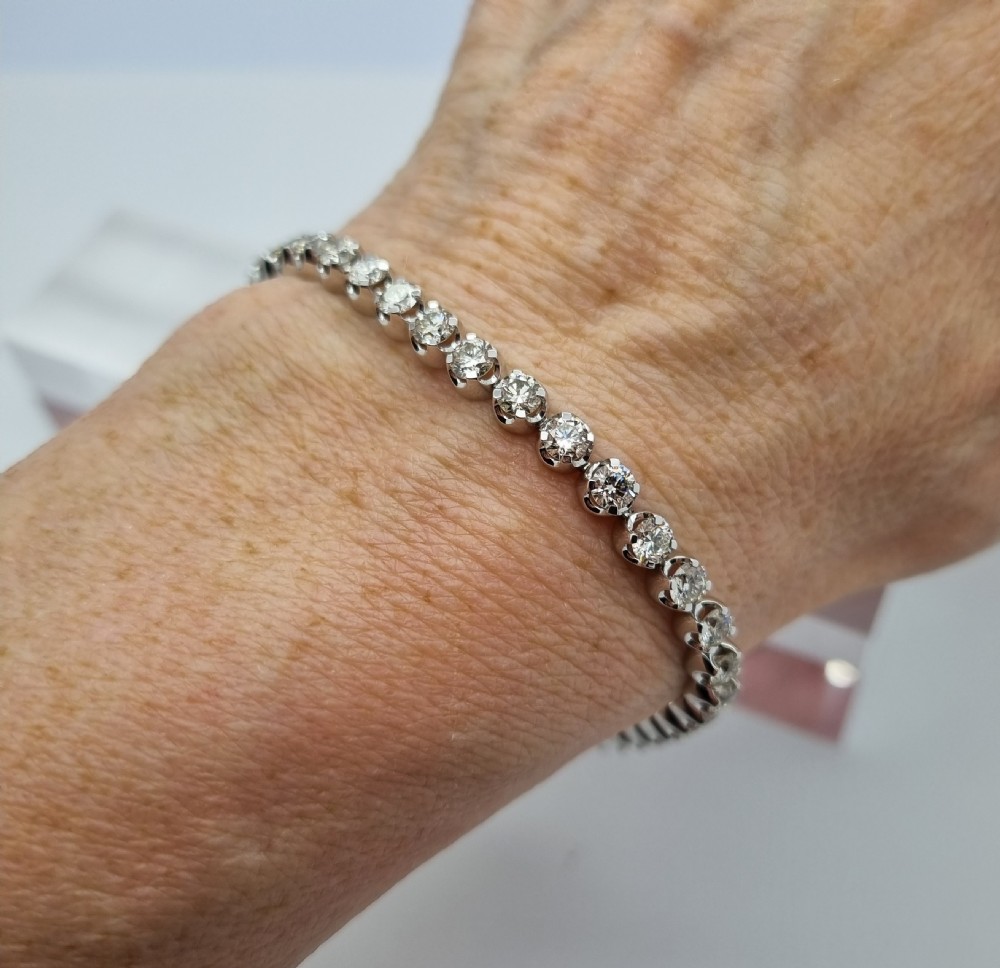 A flexible collet set diamond tennis bracelet crafted in white metal with a continuous row of round brilliant cut diamonds. View XII