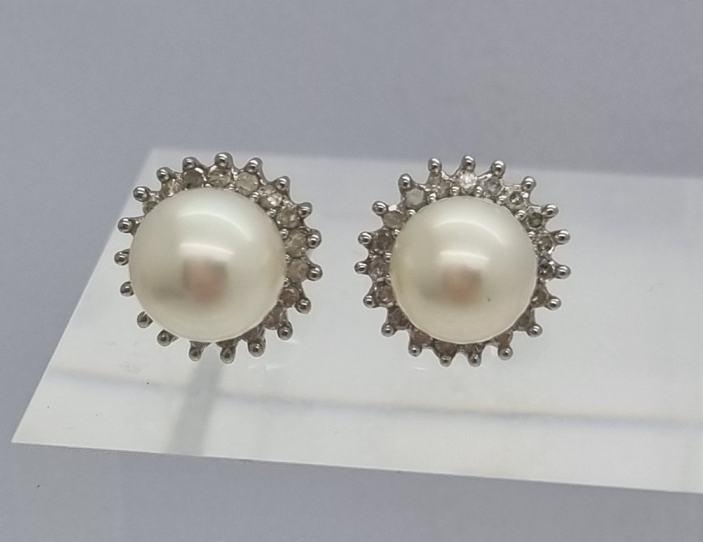 A pair of 9ct white gold stud earrings featuring round cream central pearls surrounded by halos of small round diamonds. View IV