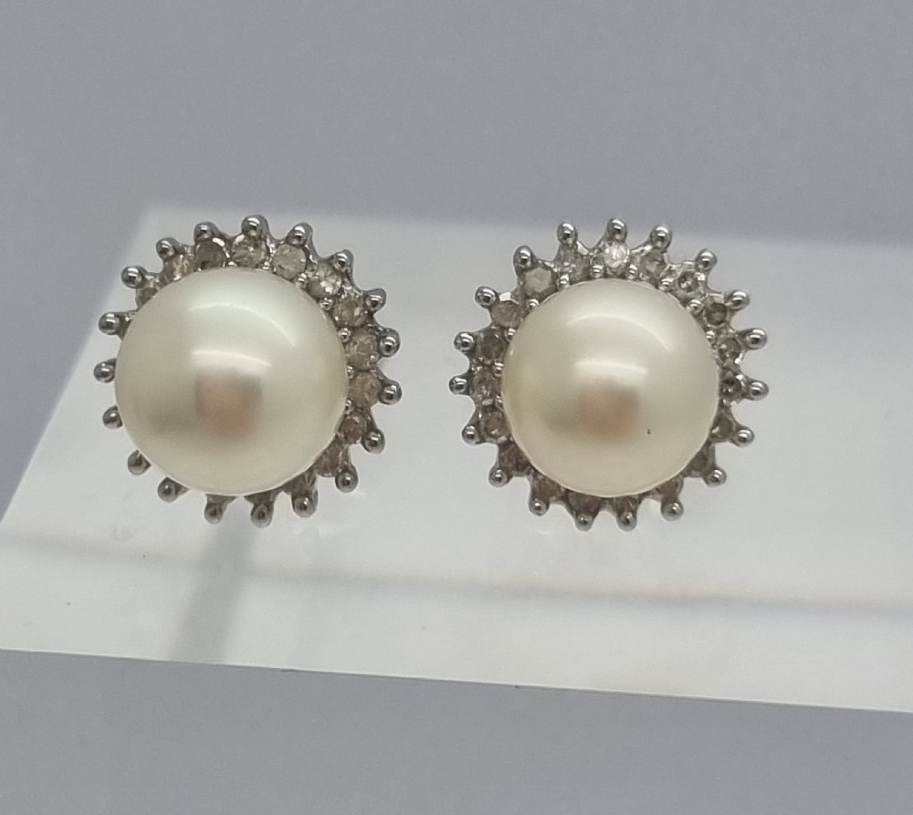 A pair of 9ct white gold stud earrings featuring round cream central pearls surrounded by halos of small round diamonds. View V