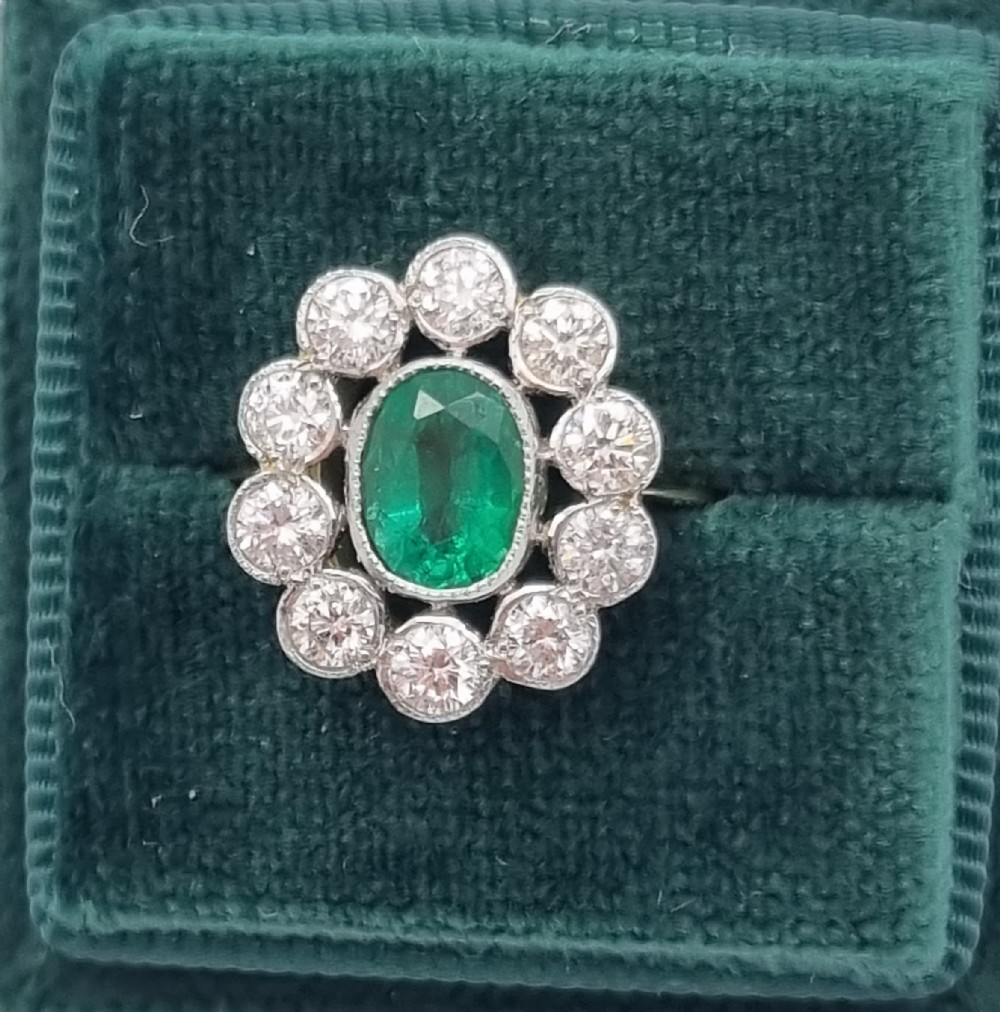 An oval-cut green emerald surrounded by a halo of ten round diamonds in a millgrained cluster setting.