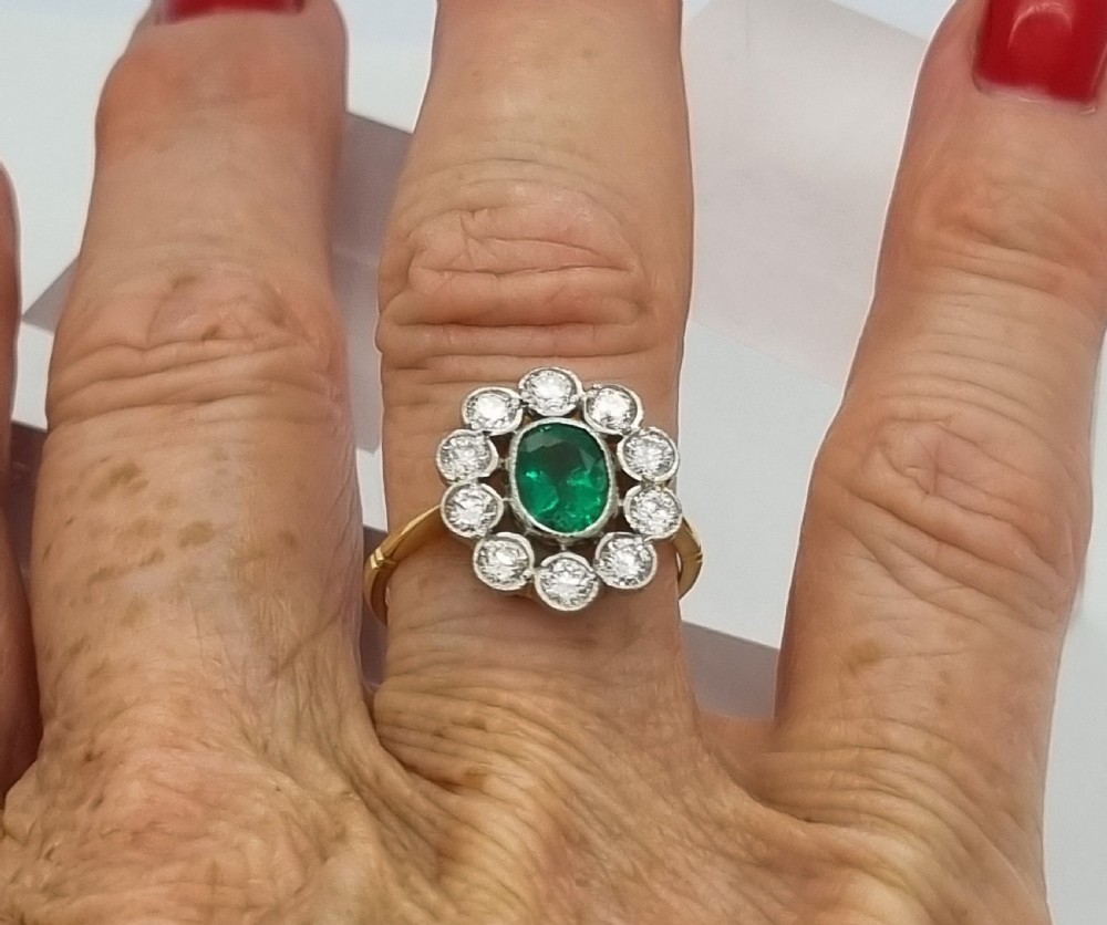 An oval-cut green emerald surrounded by a halo of ten round diamonds in a millgrained cluster setting. View II
