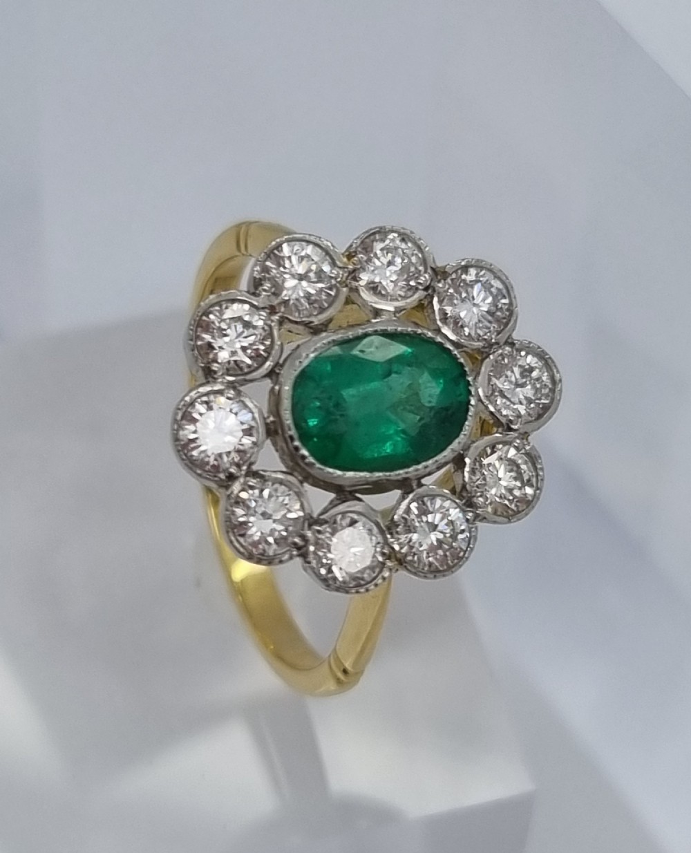 An oval-cut green emerald surrounded by a halo of ten round diamonds in a millgrained cluster setting. View VI