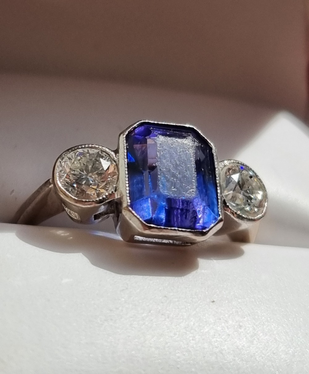 An 18ct white gold ring featuring a central emerald cut blue-violet tanzanite flanked by two round diamonds in bezel settings.
