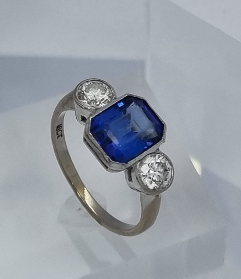 An 18ct white gold ring featuring a central emerald cut blue-violet tanzanite flanked by two round diamonds in bezel settings. View II