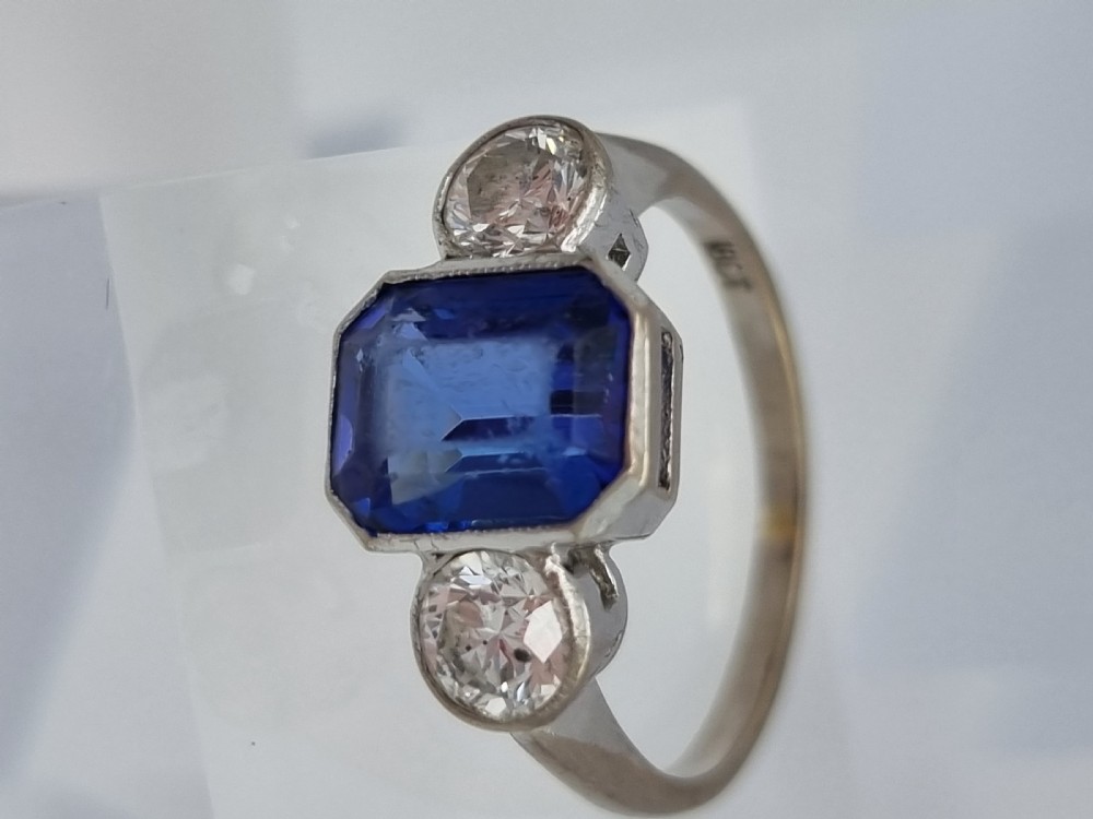 An 18ct white gold ring featuring a central emerald cut blue-violet tanzanite flanked by two round diamonds in bezel settings. View III