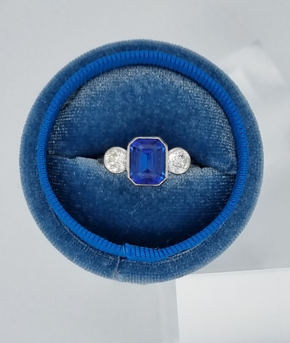 An 18ct white gold ring featuring a central emerald cut blue-violet tanzanite flanked by two round diamonds in bezel settings. View IV