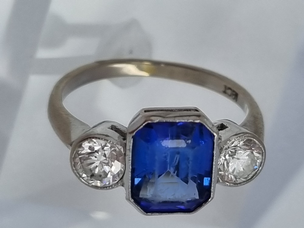 An 18ct white gold ring featuring a central emerald cut blue-violet tanzanite flanked by two round diamonds in bezel settings. View V