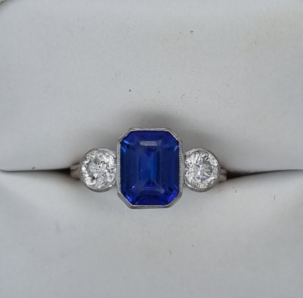 An 18ct white gold ring featuring a central emerald cut blue-violet tanzanite flanked by two round diamonds in bezel settings. View VI
