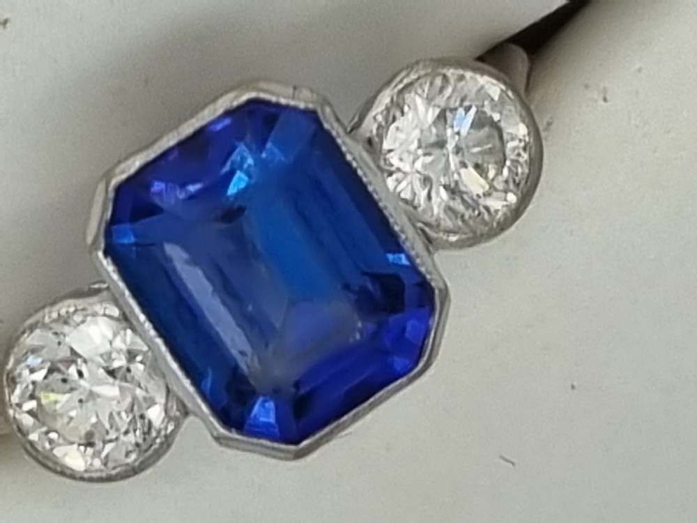 An 18ct white gold ring featuring a central emerald cut blue-violet tanzanite flanked by two round diamonds in bezel settings. View VII