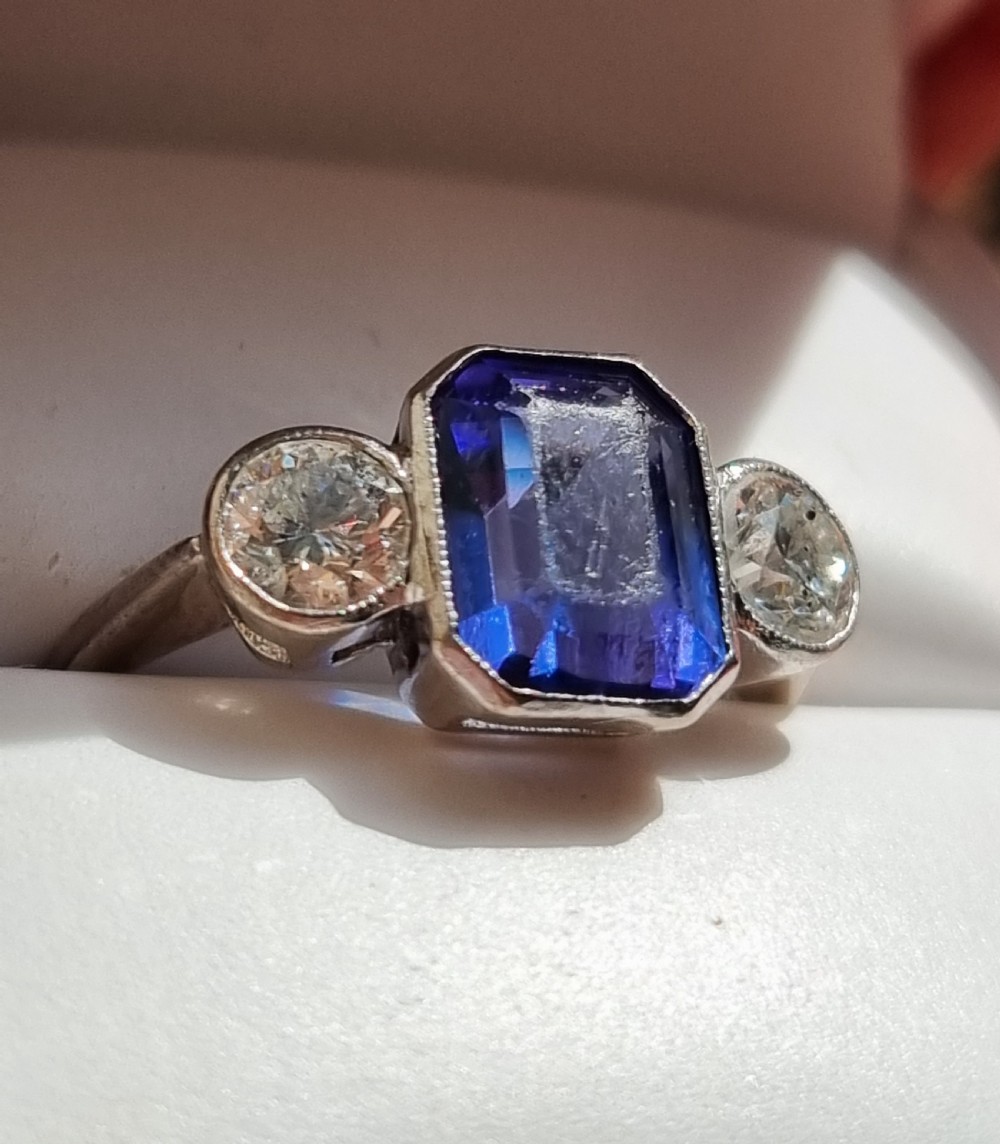 An 18ct white gold ring featuring a central emerald cut blue-violet tanzanite flanked by two round diamonds in bezel settings. View IX