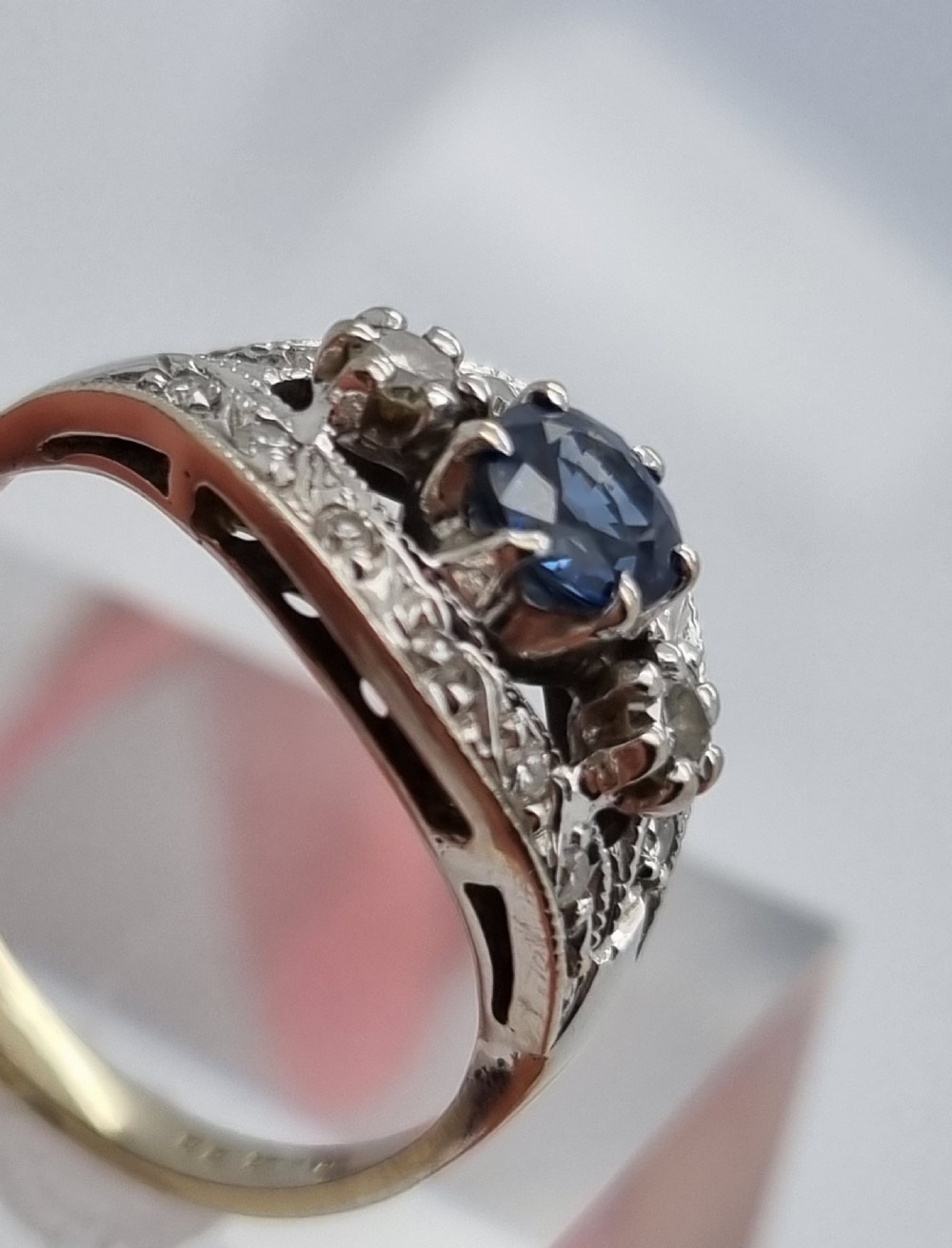 A 14ct gold dress ring featuring a central round blue sapphire surrounded by accent diamonds set in detailed milgrain white gold openwork. View IX