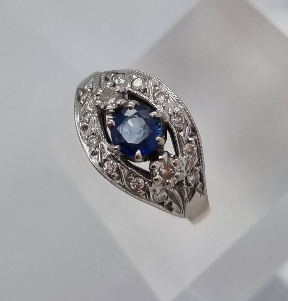 A 14ct gold dress ring featuring a central round blue sapphire surrounded by accent diamonds set in detailed milgrain white gold openwork. View X