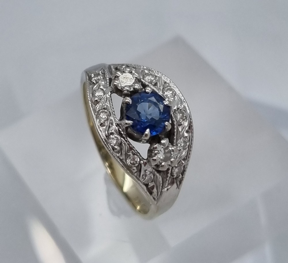 A 14ct gold dress ring featuring a central round blue sapphire surrounded by accent diamonds set in detailed milgrain white gold openwork. View XI