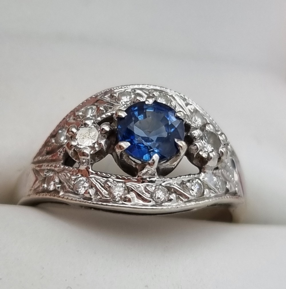 A 14ct gold dress ring featuring a central round blue sapphire surrounded by accent diamonds set in detailed milgrain white gold openwork. View XV