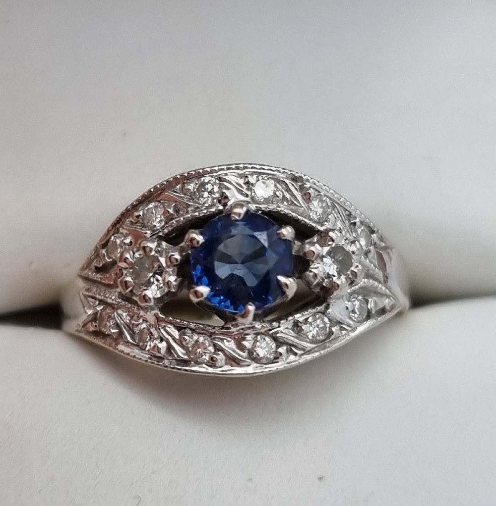 A 14ct gold dress ring featuring a central round blue sapphire surrounded by accent diamonds set in detailed milgrain white gold openwork. View XVI