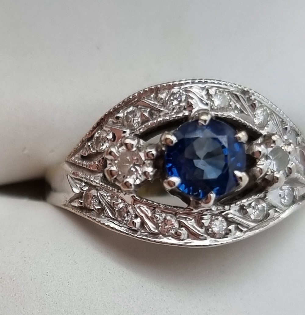 A 14ct gold dress ring featuring a central round blue sapphire surrounded by accent diamonds set in detailed milgrain white gold openwork. View XVII