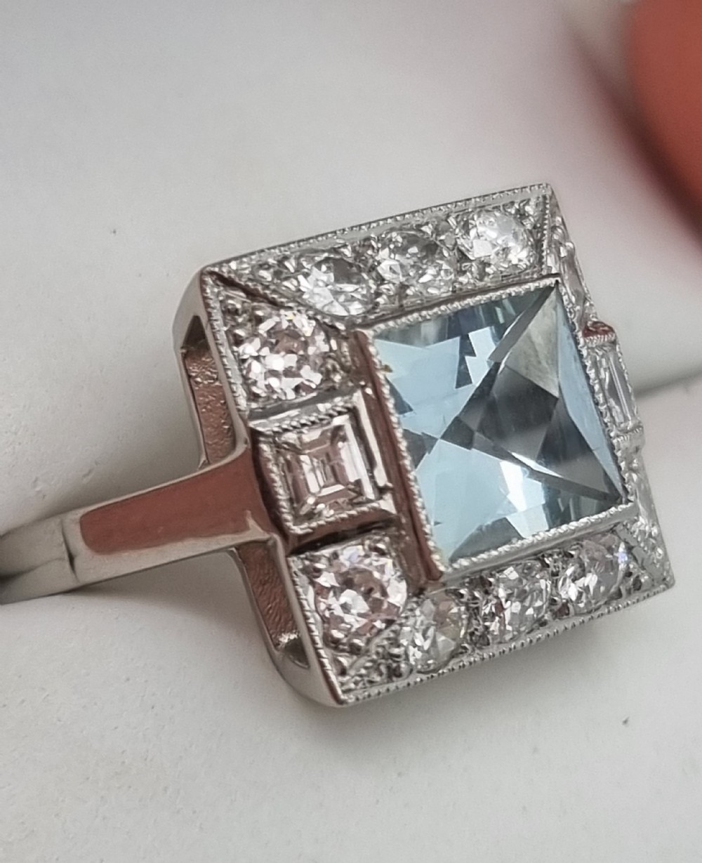 An Art Deco style platinum ring featuring a square central light blue aquamarine surrounded by a geometric border of diamonds with millegrain edging.