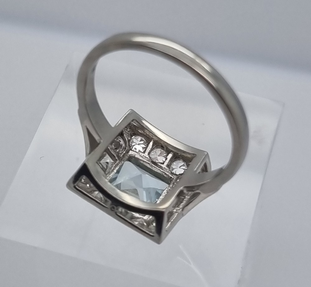 An Art Deco style platinum ring featuring a square central light blue aquamarine surrounded by a geometric border of diamonds with millegrain edging. View VI