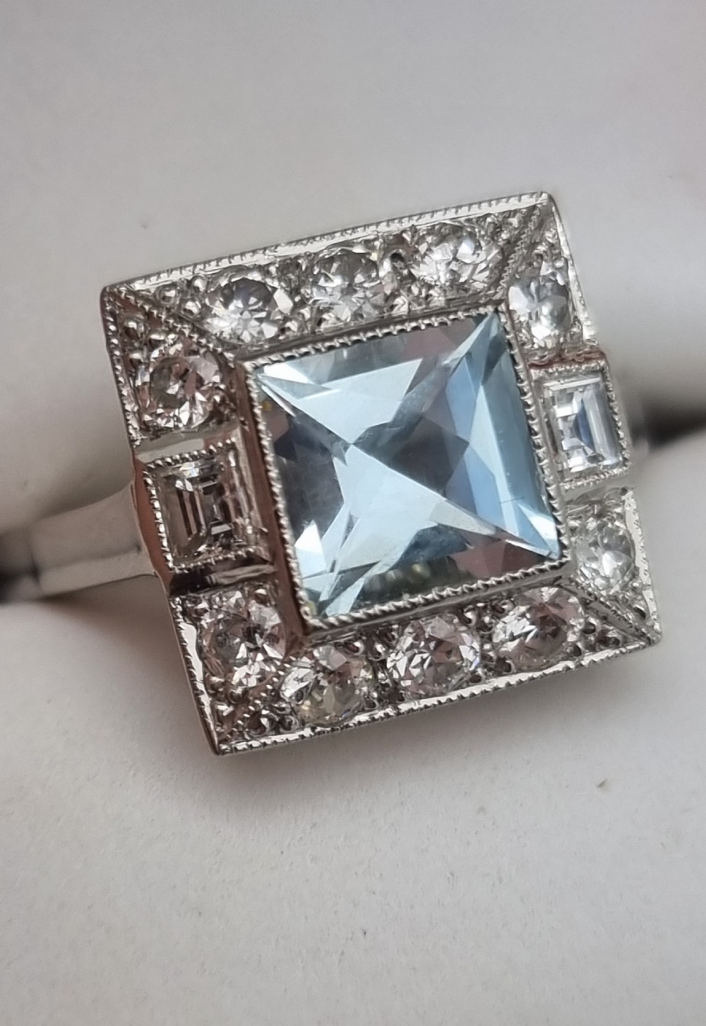 An Art Deco style platinum ring featuring a square central light blue aquamarine surrounded by a geometric border of diamonds with millegrain edging. View VIII