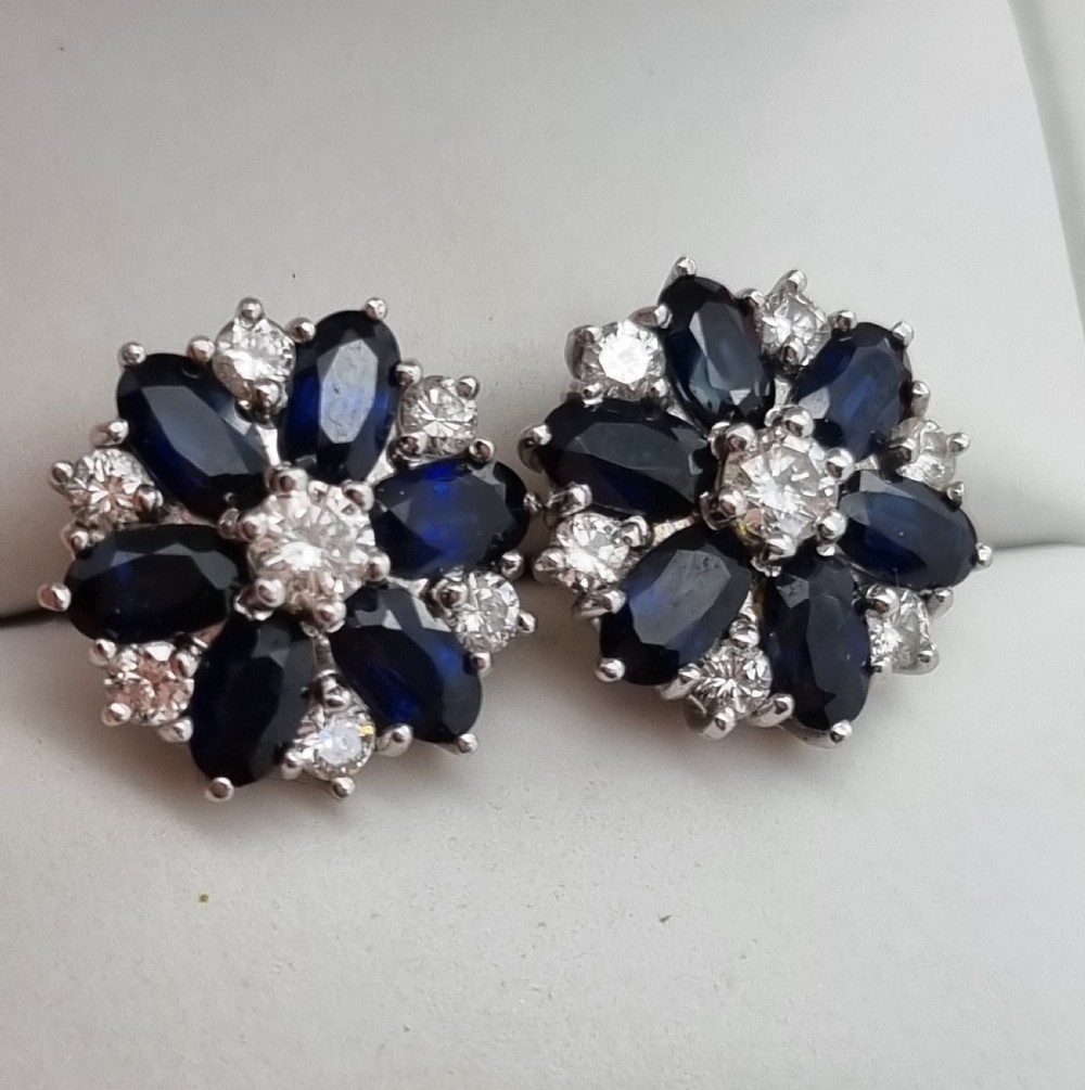 A pair of cluster stud earrings featuring deep blue sapphires arranged in a floral motif around a central diamond, set in white metal.