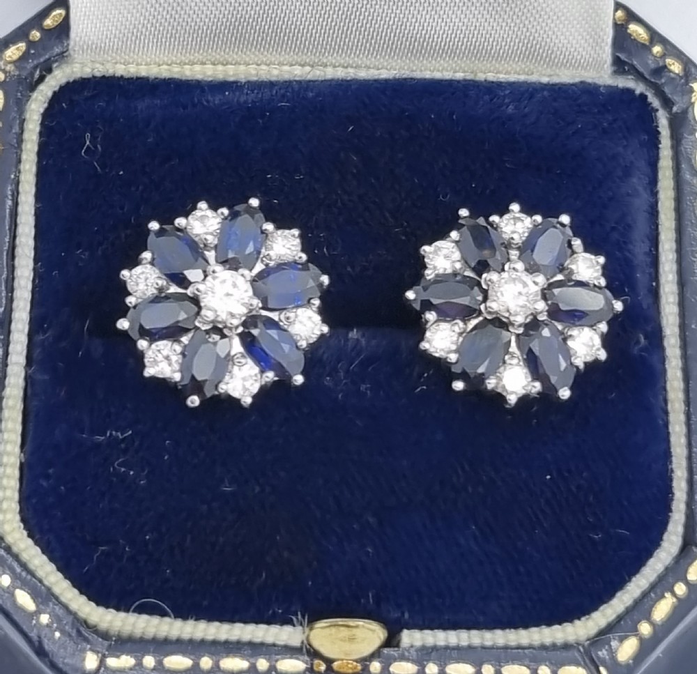 A pair of cluster stud earrings featuring deep blue sapphires arranged in a floral motif around a central diamond, set in white metal. View II