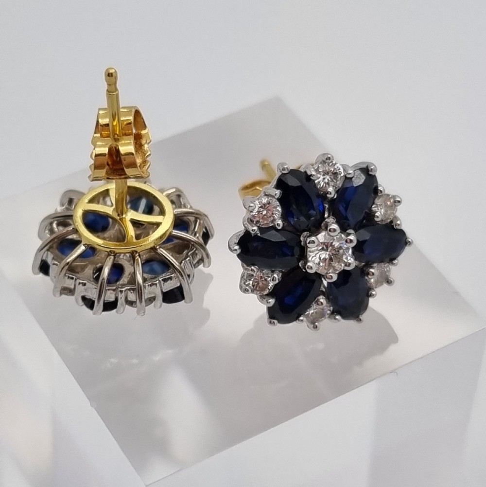 A pair of cluster stud earrings featuring deep blue sapphires arranged in a floral motif around a central diamond, set in white metal. View IV
