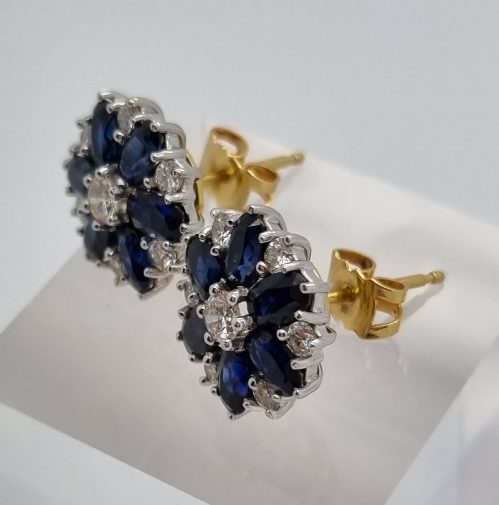 A pair of cluster stud earrings featuring deep blue sapphires arranged in a floral motif around a central diamond, set in white metal. View V
