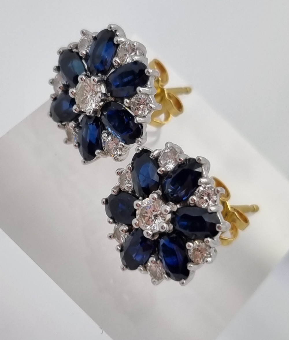 A pair of cluster stud earrings featuring deep blue sapphires arranged in a floral motif around a central diamond, set in white metal. View VI