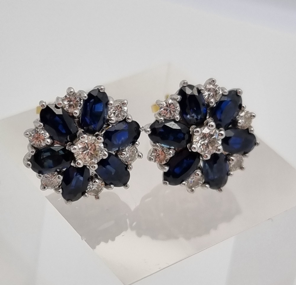 A pair of cluster stud earrings featuring deep blue sapphires arranged in a floral motif around a central diamond, set in white metal. View VII
