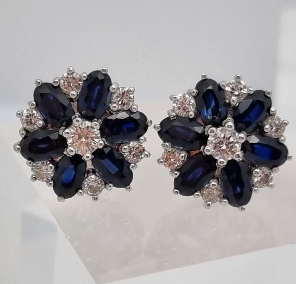 A pair of cluster stud earrings featuring deep blue sapphires arranged in a floral motif around a central diamond, set in white metal. View VIII