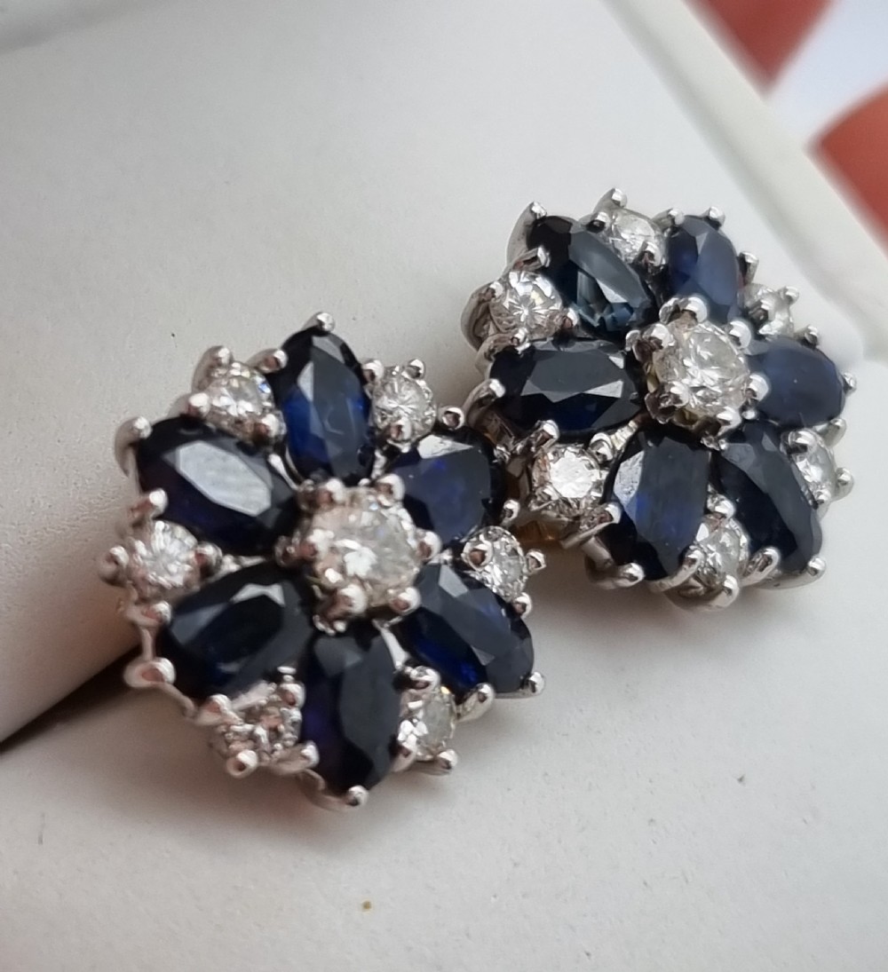A pair of cluster stud earrings featuring deep blue sapphires arranged in a floral motif around a central diamond, set in white metal. View IX