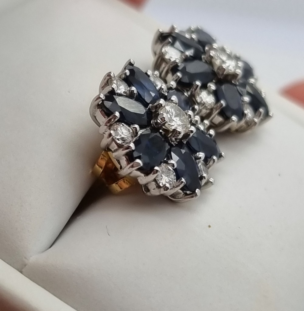 A pair of cluster stud earrings featuring deep blue sapphires arranged in a floral motif around a central diamond, set in white metal. View X