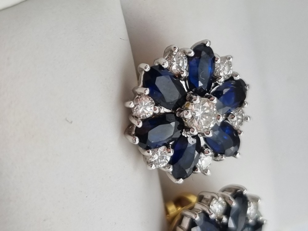 A pair of cluster stud earrings featuring deep blue sapphires arranged in a floral motif around a central diamond, set in white metal. View XIII