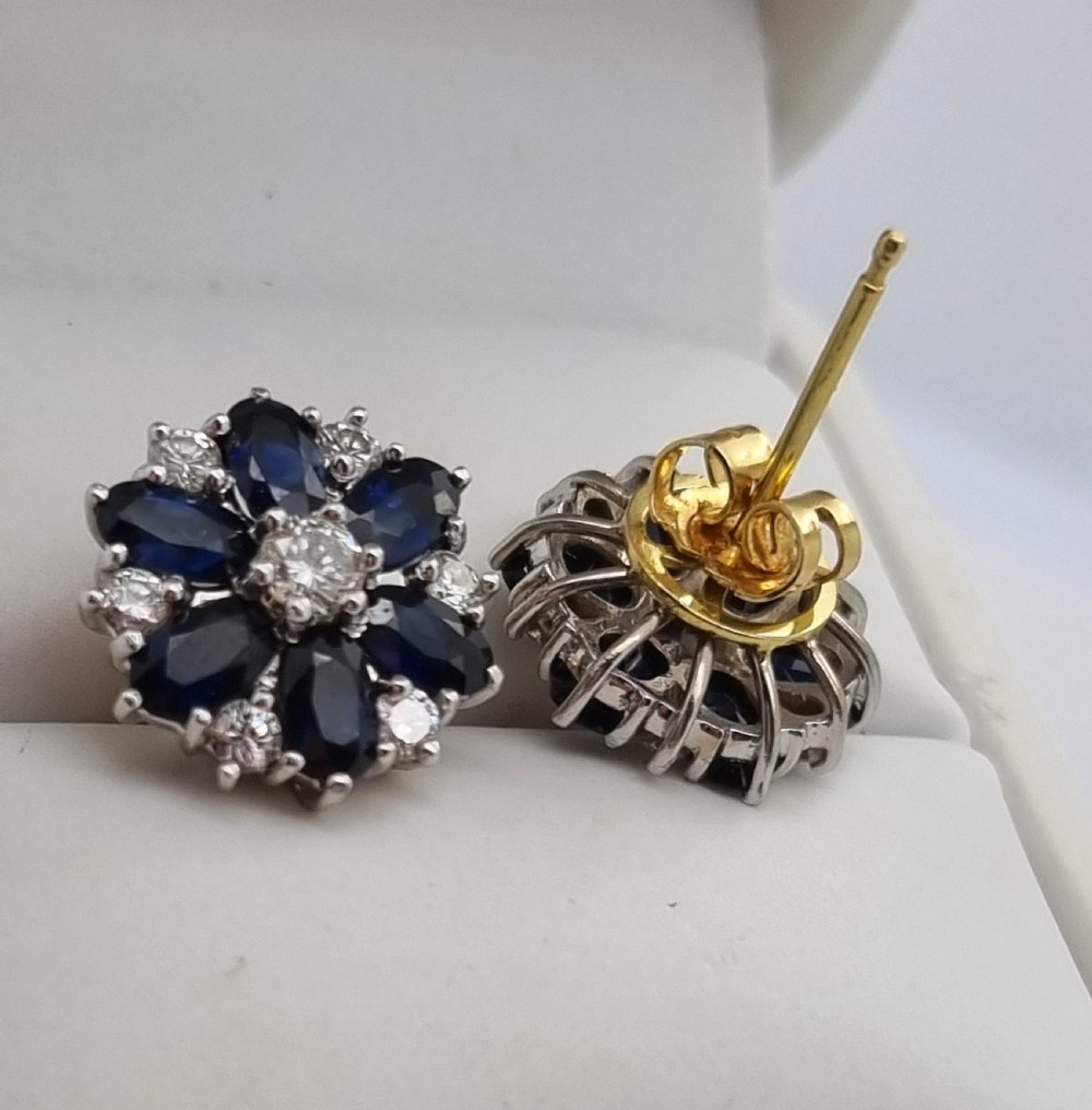 A pair of cluster stud earrings featuring deep blue sapphires arranged in a floral motif around a central diamond, set in white metal. View XIV