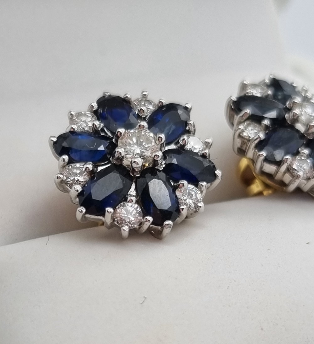 A pair of cluster stud earrings featuring deep blue sapphires arranged in a floral motif around a central diamond, set in white metal. View XV