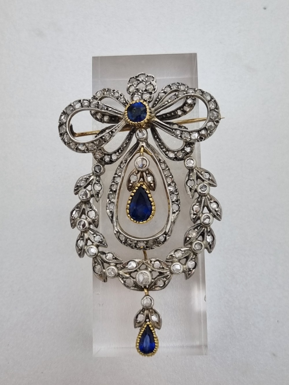 An Edwardian bow and garland design brooch pendant crafted in silver-topped gold, set with diamonds and teardrop blue sapphires. View II