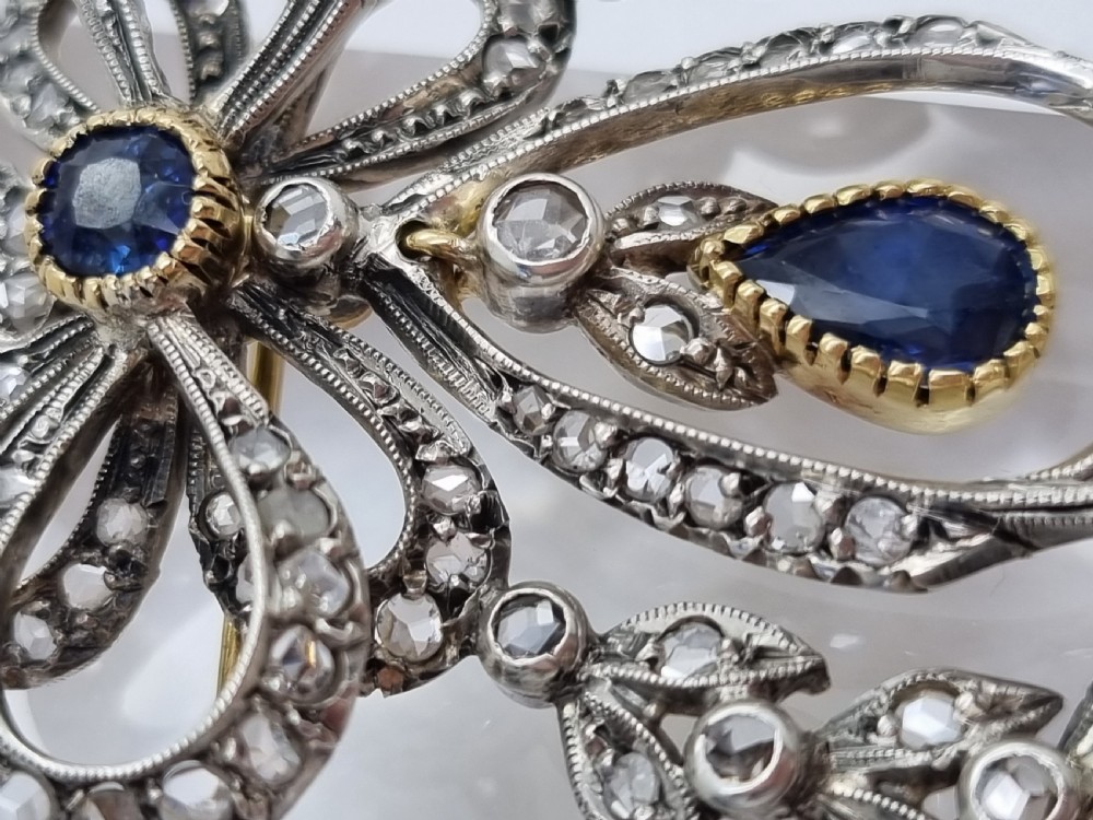 An Edwardian bow and garland design brooch pendant crafted in silver-topped gold, set with diamonds and teardrop blue sapphires. View IV