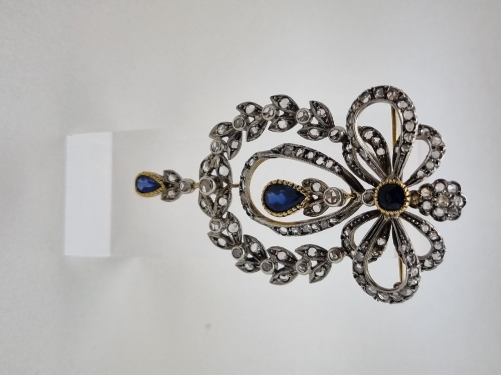 An Edwardian bow and garland design brooch pendant crafted in silver-topped gold, set with diamonds and teardrop blue sapphires. View V
