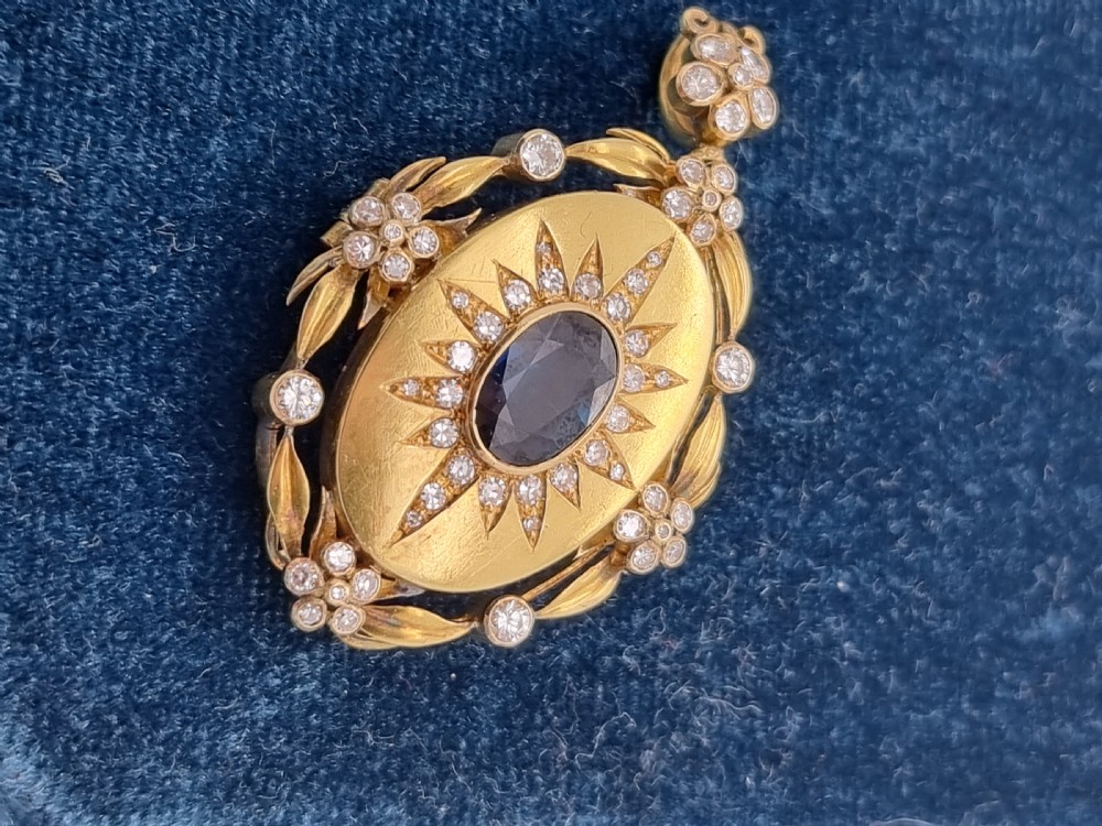 An oval yellow gold Victorian pendant brooch featuring a central dark blue sapphire framed within a starburst of small diamonds and surround of foliate motifs.