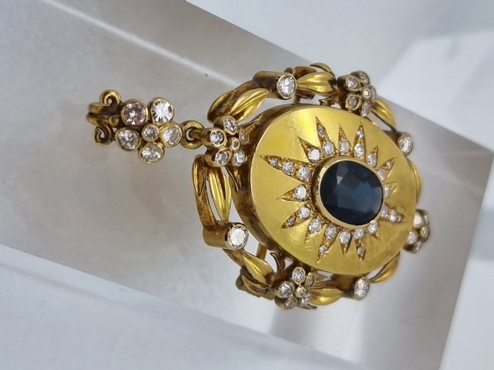 An oval yellow gold Victorian pendant brooch featuring a central dark blue sapphire framed within a starburst of small diamonds and surround of foliate motifs. View II
