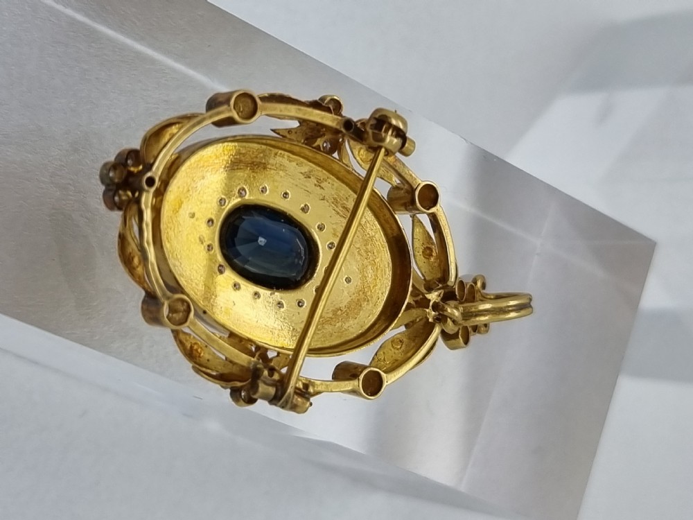 An oval yellow gold Victorian pendant brooch featuring a central dark blue sapphire framed within a starburst of small diamonds and surround of foliate motifs. View III