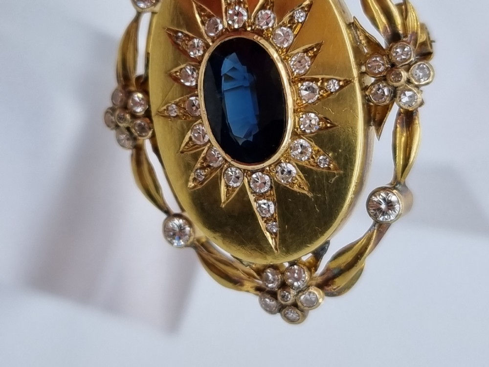 An oval yellow gold Victorian pendant brooch featuring a central dark blue sapphire framed within a starburst of small diamonds and surround of foliate motifs. View V