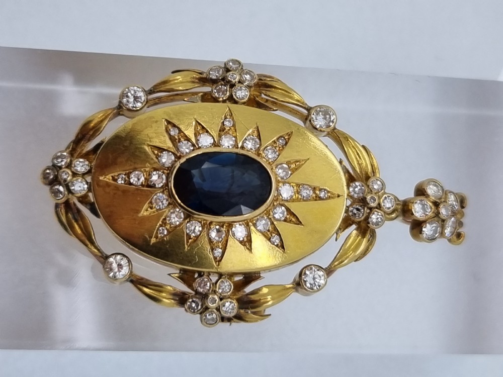 An oval yellow gold Victorian pendant brooch featuring a central dark blue sapphire framed within a starburst of small diamonds and surround of foliate motifs. View VI