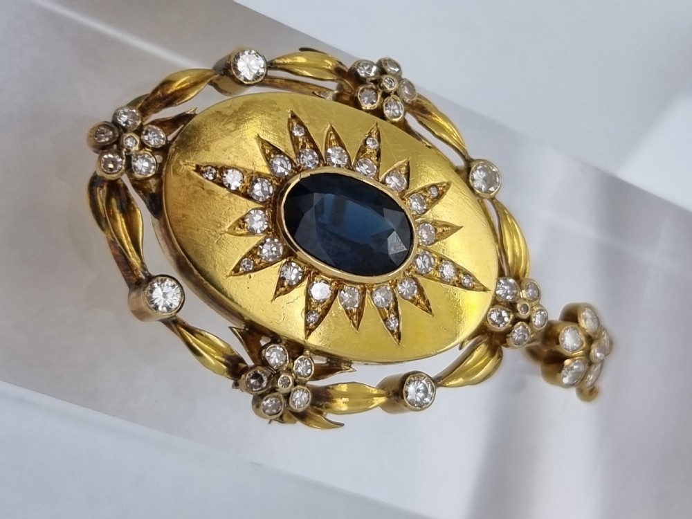 An oval yellow gold Victorian pendant brooch featuring a central dark blue sapphire framed within a starburst of small diamonds and surround of foliate motifs. View VII