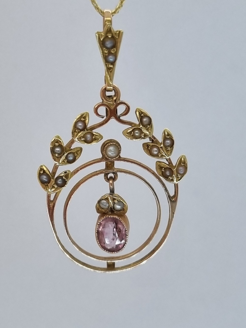 An Edwardian 15ct gold pendant featuring seed pearl leaf motifs, concentric gold rings, and a central suspended oval pink gemstone.