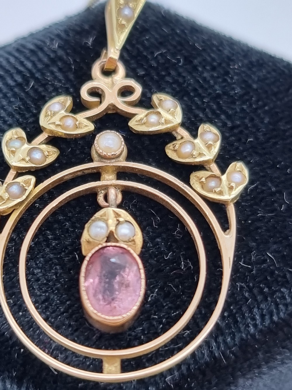 An Edwardian 15ct gold pendant featuring seed pearl leaf motifs, concentric gold rings, and a central suspended oval pink gemstone. View V