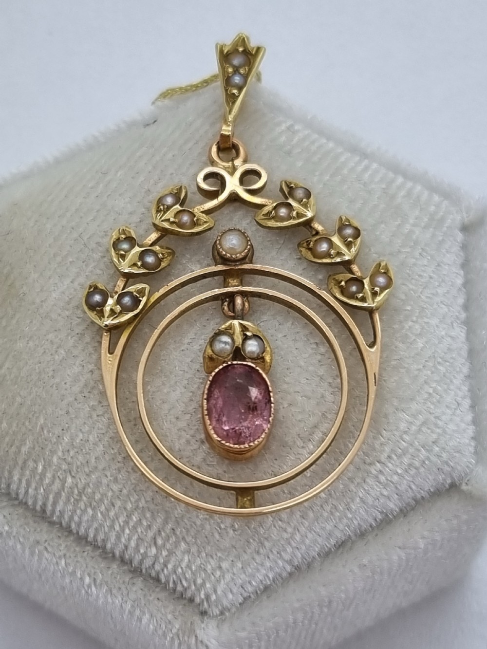 An Edwardian 15ct gold pendant featuring seed pearl leaf motifs, concentric gold rings, and a central suspended oval pink gemstone. View VI