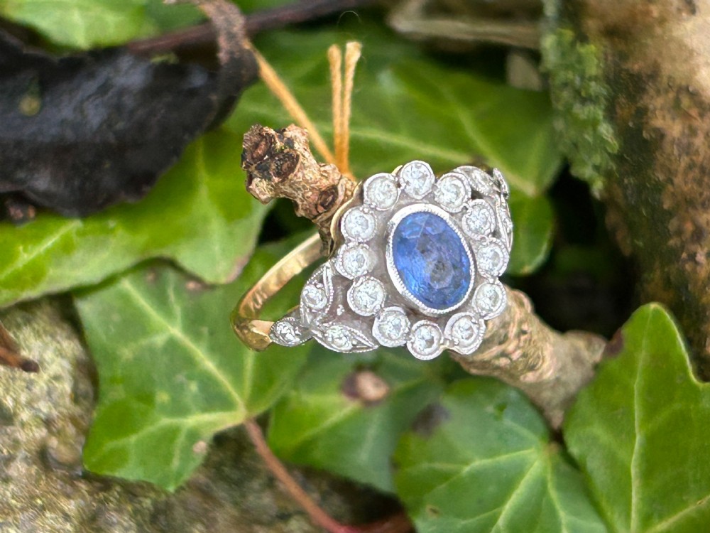 An Edwardian oval blue sapphire cluster ring surrounded by a diamond halo with foliate shoulders and a yellow gold shank. View II