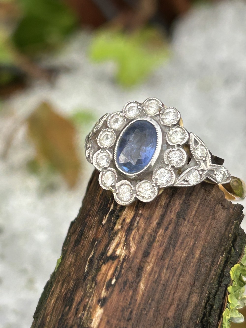 An Edwardian oval blue sapphire cluster ring surrounded by a diamond halo with foliate shoulders and a yellow gold shank.